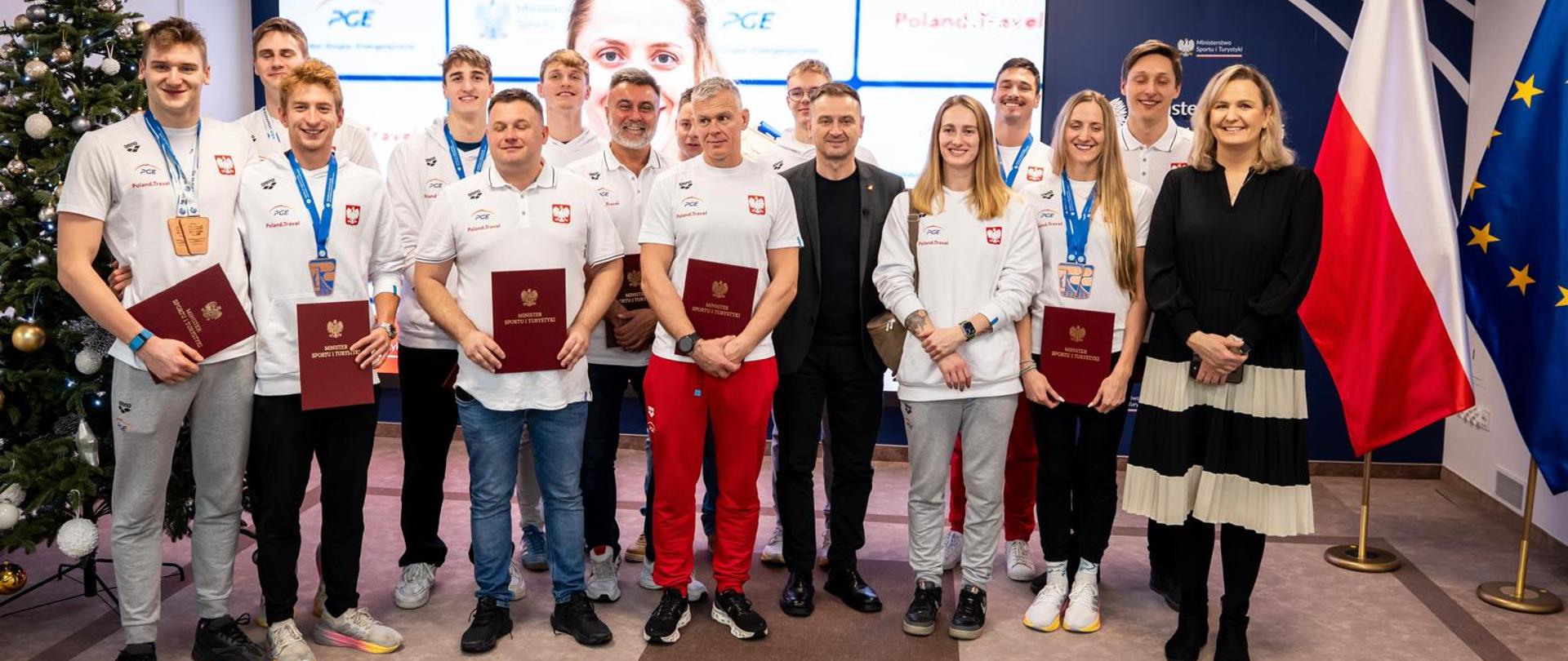 Zdjęcie zbiorowe, a na nim Minister Sportu i Turystyki Sławomir Nitras, pływacy Ksawery Masiuk, Kacper Stokowski, Piotr Ludwiczak, Krzysztof Chmielewski, Katarzyna Wasick, Jakub Majerski, Kamil Sieradzki, Dominika Sztandera, Adrian Jaśkiewicz, a także trenerzy Paweł Wołkow, Grzegorz Widanka, Dominik Grudziński, Jakub Karpiński i Robert Wilk oraz prezes Polskiego Związku Pływackiego Otylia Jędrzejczak. Za uczestnikami spotkania granatowa ścianka z logotypami MSiT, na niej duży ekran. Z prawej strony zdjęcia choinka, z lewej - flagi Polski i UE.