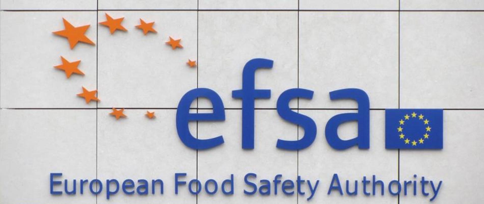 Niebieski napis efsa. Wyżej po lewej stronie 9 gwiazdek w pomarańczowym kolorze. Po prawej od napisu efsa flaga unii europejskiej. Pod napisem efsa rozwinięcie skrótu European Food Safety Authority