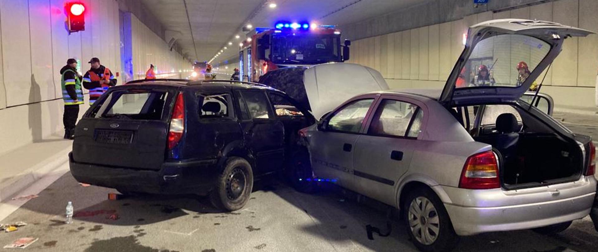 Dwa samochody po wypadku, jeden szary drugi czarny. Naokoło rozrzucone są części samochodów. W tle samochód strażacki. Wszystko dzieje się w tunelu.