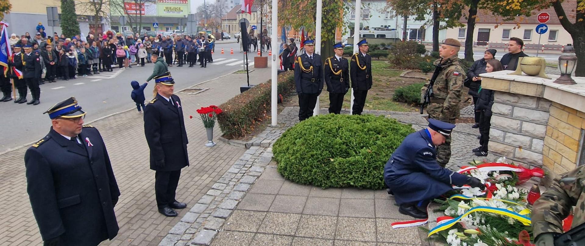 Przy pomniku Komendant Powiatowy Policji składa wieniec. Przed podwyższeniem stoją oficerowie PSP. W tle trzy białe maszty z polskimi flagami. Przed nimi stoi poczet flagowy strażaków PSP. Obok pomnika warta pełniona przez żołnierza WP. W dalszym tle zgromadzeni uczestnicy uroczystości. 