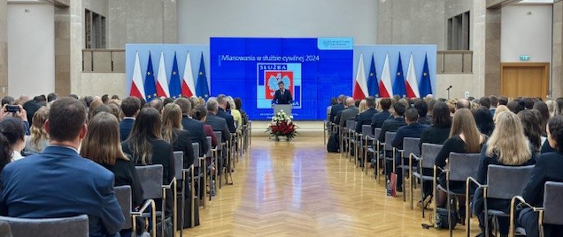 Sala konferencyjna i. A. Walentynowicz w gmachu KPRM, w tle logo służby cywilnej i flagi Polski oraz UE. Przy mównicy mężczyzna - szef Kancelarii PRM, Jan Grabiec, po obu stronach na krzesłach siedzą uczestnicy uroczystości - nowo mianowani urzędnicy i zaproszeni goście.