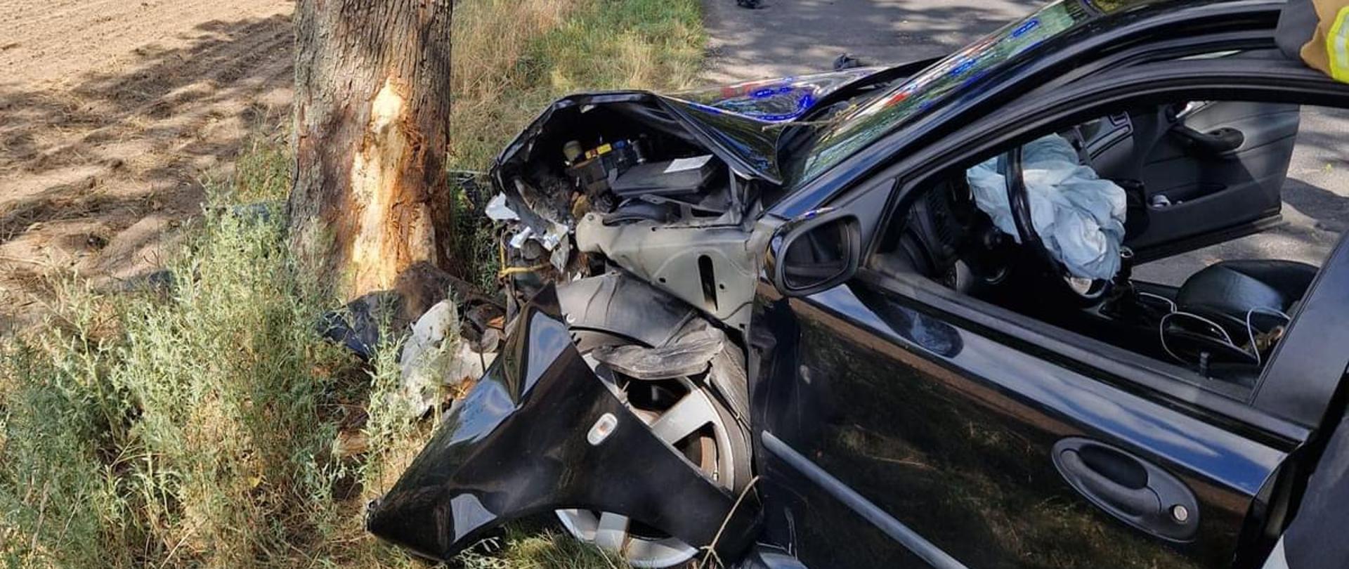 Samochód osobowy koloru ciemnego po uderzeniu w przydrożne drzewo. W tle na drodze stoi wóz strażacki.