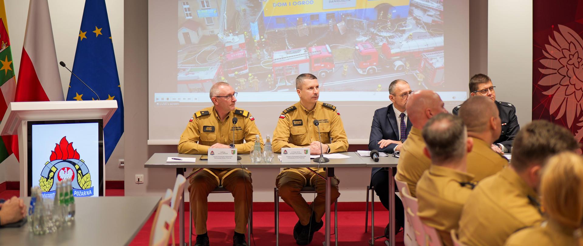 Panel dyskusyjny w trakcie odprawy rocznej. Przy stole prezydialnym siedzi kilku strażaków w mundurach, w tym zastępca komendanta głównego, oraz przedstawiciele administracji. Na ekranie w tle widoczny jest slajd z obrazem przedstawiającym akcję gaśniczą budynku handlowego, opatrzony napisem „Dom i Ogród”. Na stole znajdują się mikrofony, butelki z wodą i tabliczki z nazwiskami uczestników panelu.