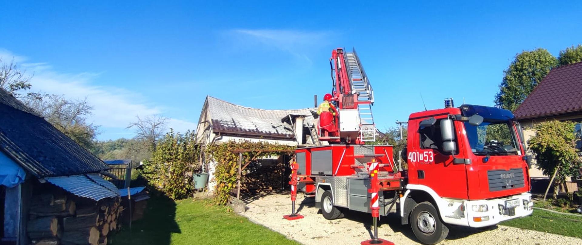 Podnośnik hydrauliczny rozłożony koloru czerwonego, po lewej stronie kawałek ułożonego drzewa, po prawej stronie dom. W tle budynek gospodarczy 