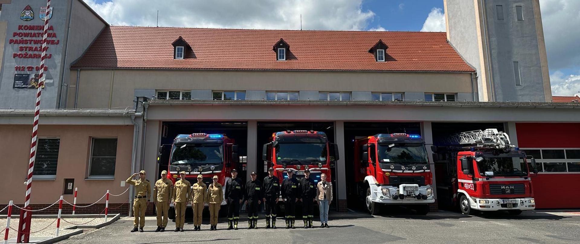 Strażacy oddali hołd tragicznie zmarłemu żołnierzowi. Na zdjęciu widać strażaków przed budynkiem Komendy, samochody strażackie wyjechały z garażu, mają włączone sygnały świetlne i dźwiękowe, widać maszt z flagą Polski i budynek Komendy Powiatowej PSP w Kościanie.