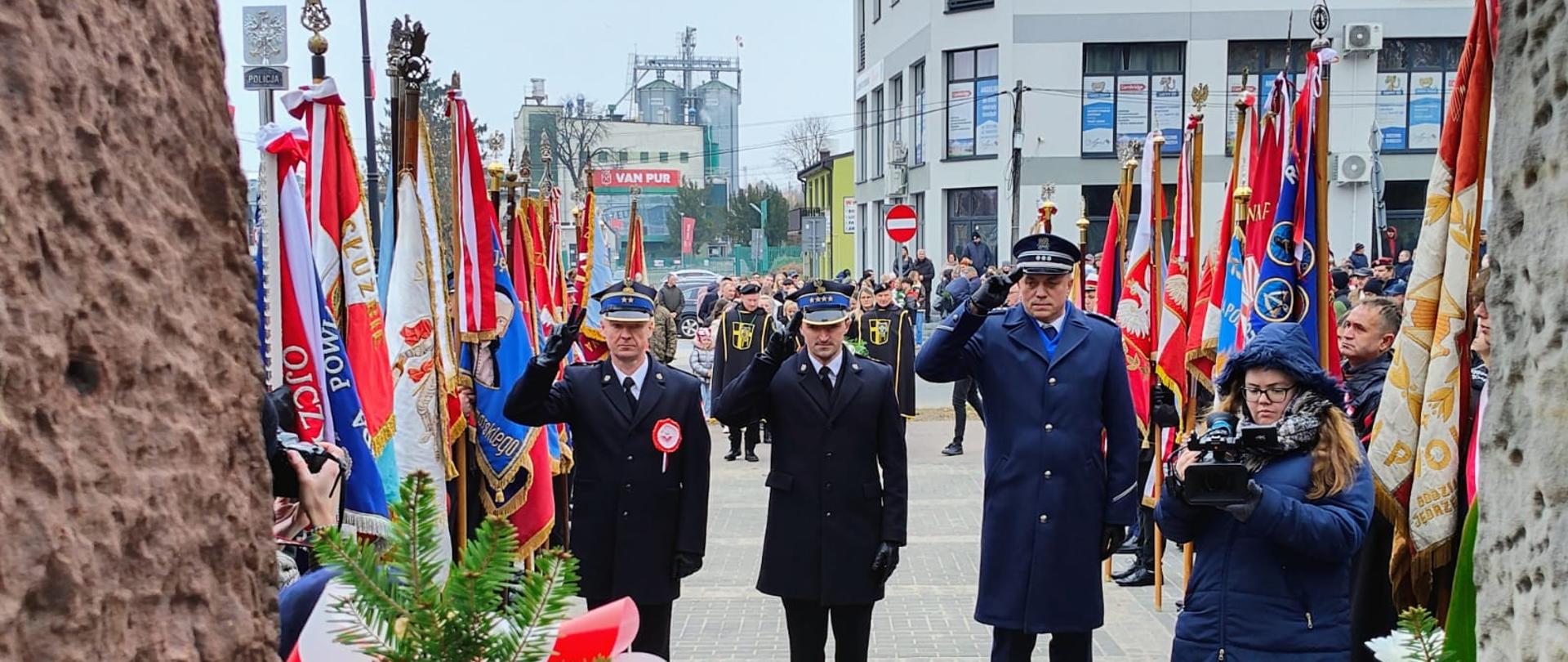 Zdjęcie obrazuje dwóch funkcjonariuszy PSP oraz Policji w umundurowaniu wyjściowym. Jeden z nich niesie wiązankę z biało-czerwonych kwiatów.