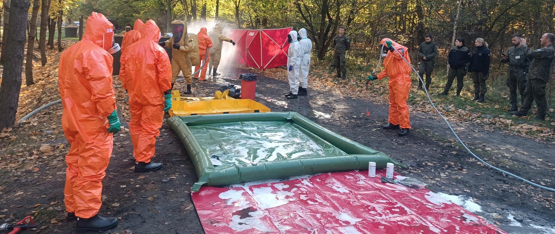 kilku ratowników chemicznych ubranych w ubrania ochrony przeciwchemicznej stoi przy prowizorycznym ciągu dekontaminacyjnych, obserwowani są przez żołnierzy