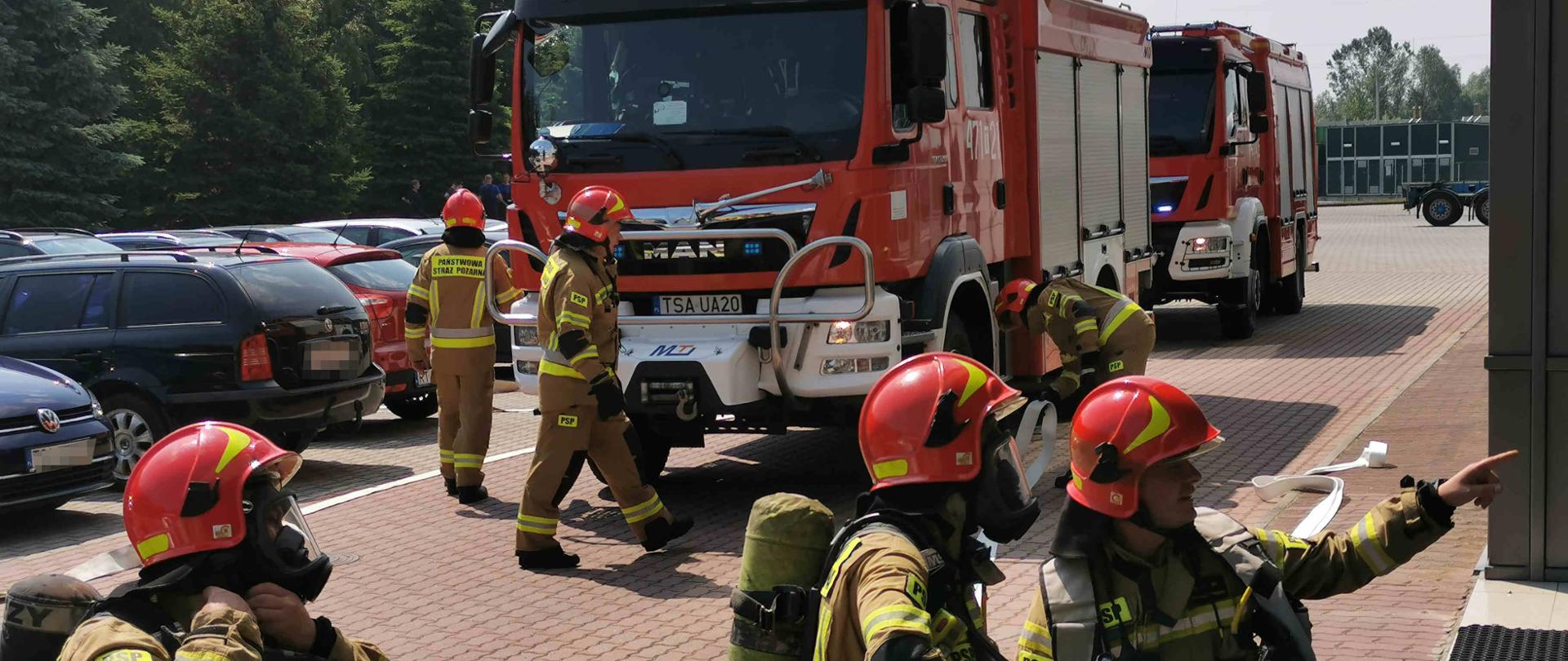 Zdjęcie zrobione w dzień. Na zdjęciu widać samochody strażackie i strażaków przygotowujących się do wejścia do budynku. 