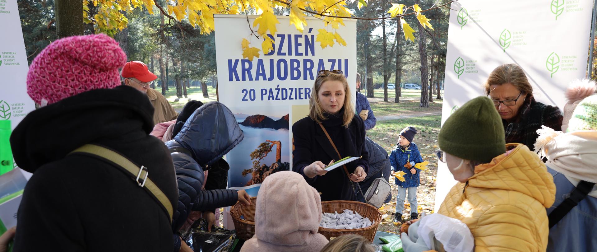 W centralnej części zdjęcia kobieta o jasnych włosach rozdaje ulotki. Przed sobą ma stół na którym znajdują się produkty reklamowe: kosze ze słodyczami, gadżety z logotypem Generalnej Dyrekcji Ochrony Środowiska. Za plecami ma roll-up z napisem Dzień Krajobrazu 20 października. Wokół niej grupka osób ubranych w kurtki i czapki (wśród nich również dzieci) wybierają dla siebie oferowane gadżety reklamowe.