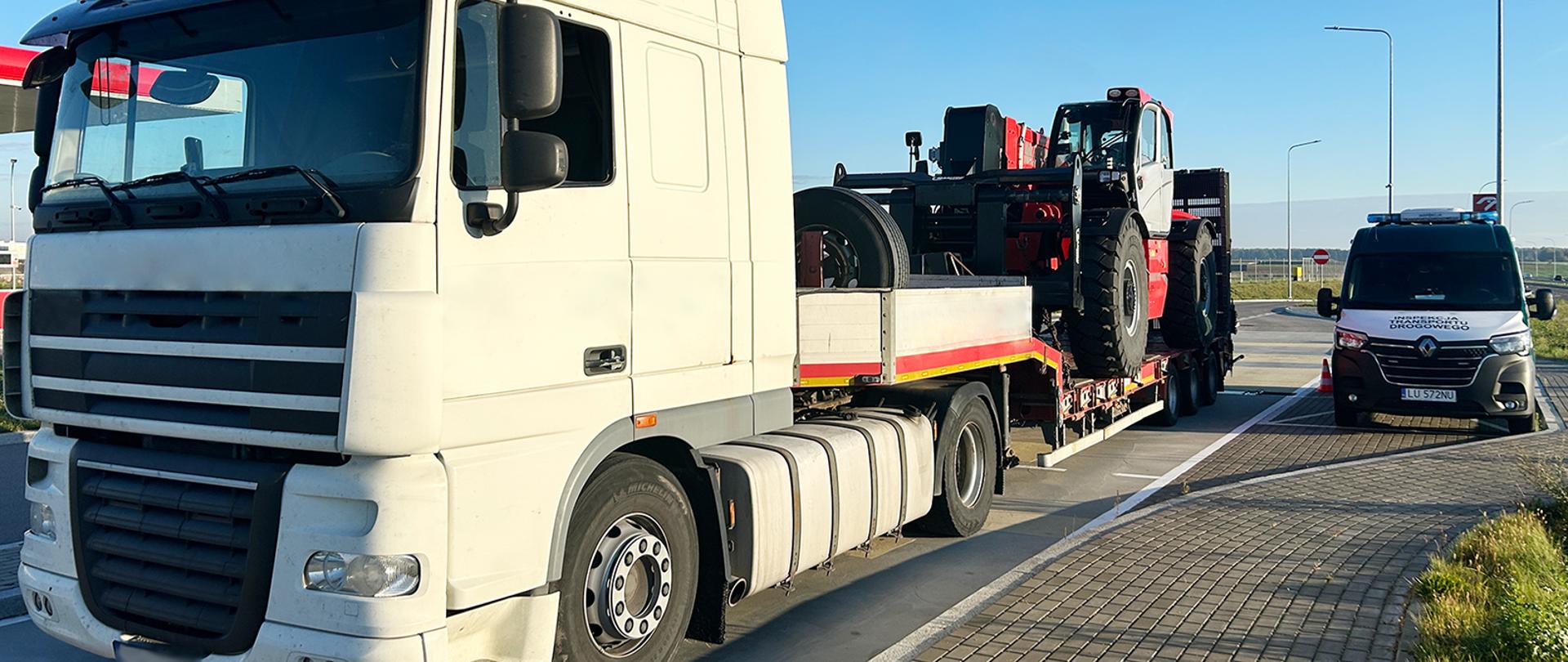 Na pierwszym planie ciągnik siodłowy z naczepą. Na na naczepie załadowana jest wielka ładowarka. Po prawej inspekcyjny furgon. Pojazdy stoją na parkingu.