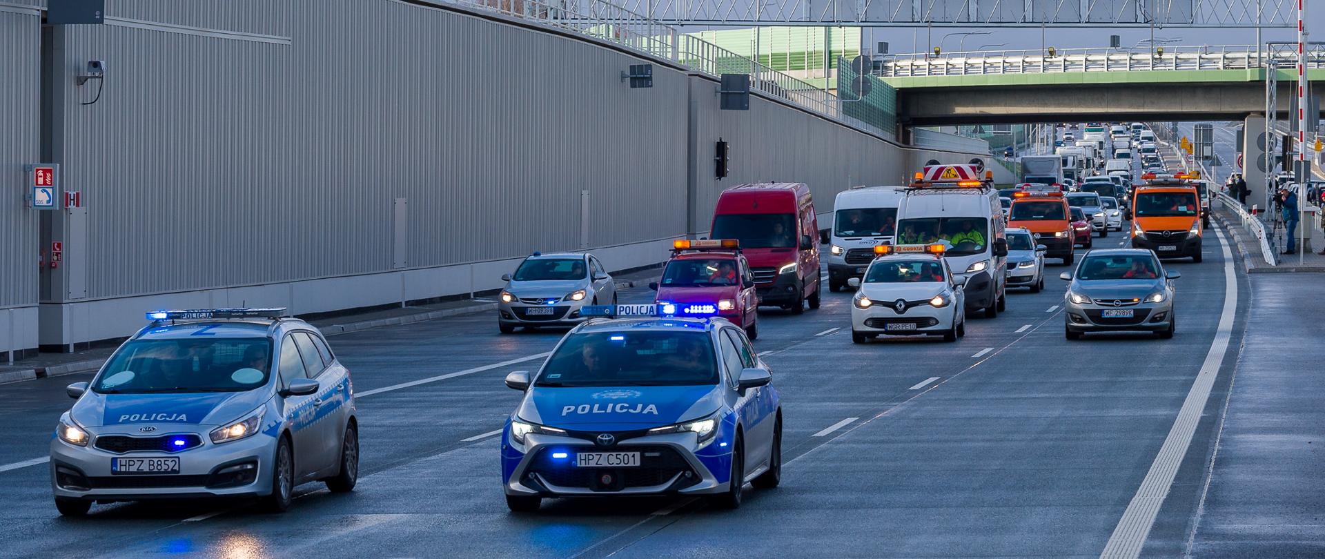 Zdjęcie przedstawia scenę na wielopasmowej drodze z wieloma pojazdami, w tym dwoma radiowozami z migającymi niebieskimi światłami, czerwonym vanem oraz kilkoma innymi samochodami i pojazdami serwisowymi GDDKiA. Wszystkie pojazdy poruszają się za dwoma radiowozami na pierwszym planie.