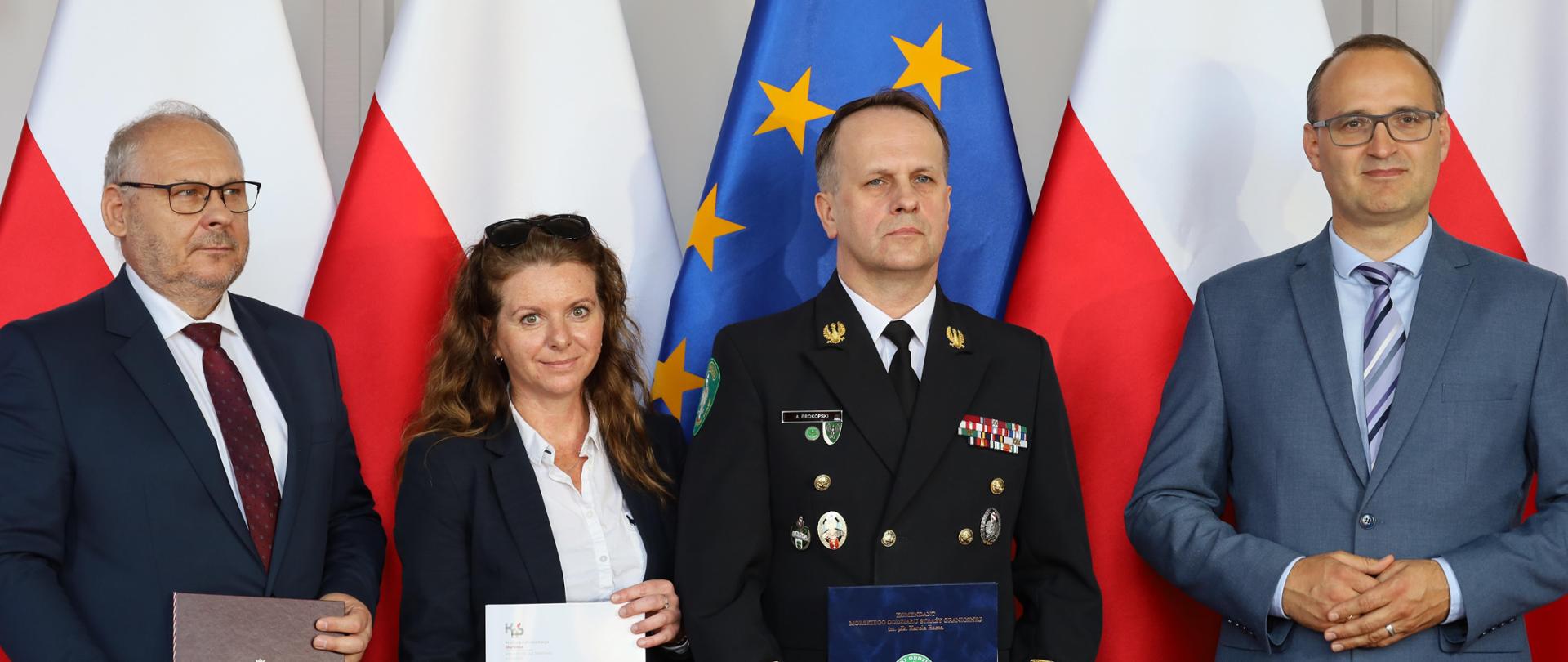 Czterech dorosłych ludzi stojących na tle flag Polski i Unii Europejskiej. Osoby na zdjęciu są ubrane formalnie, a troje z nich trzyma dokument.