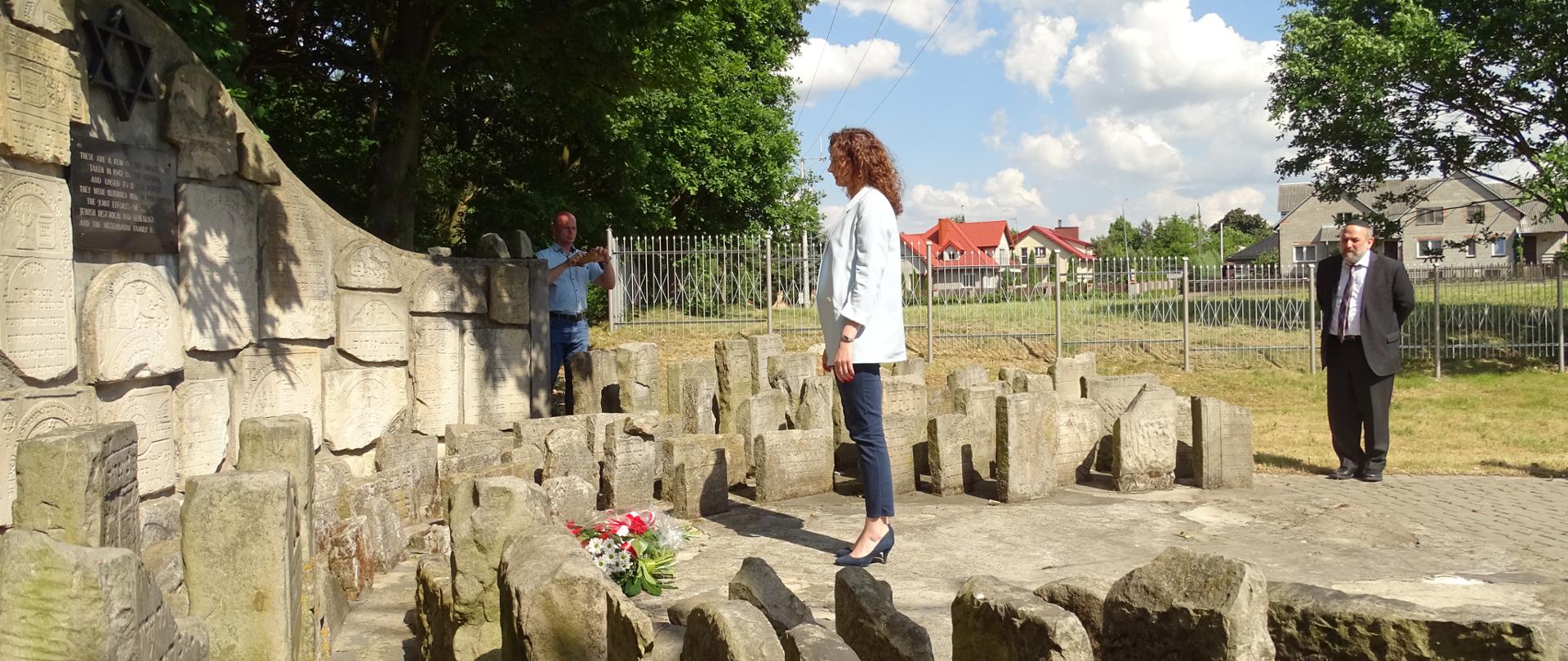 Na zdjęciu znajduje się kobieta i dwóch mężczyzn w tle na cmentarzu żydowskim 