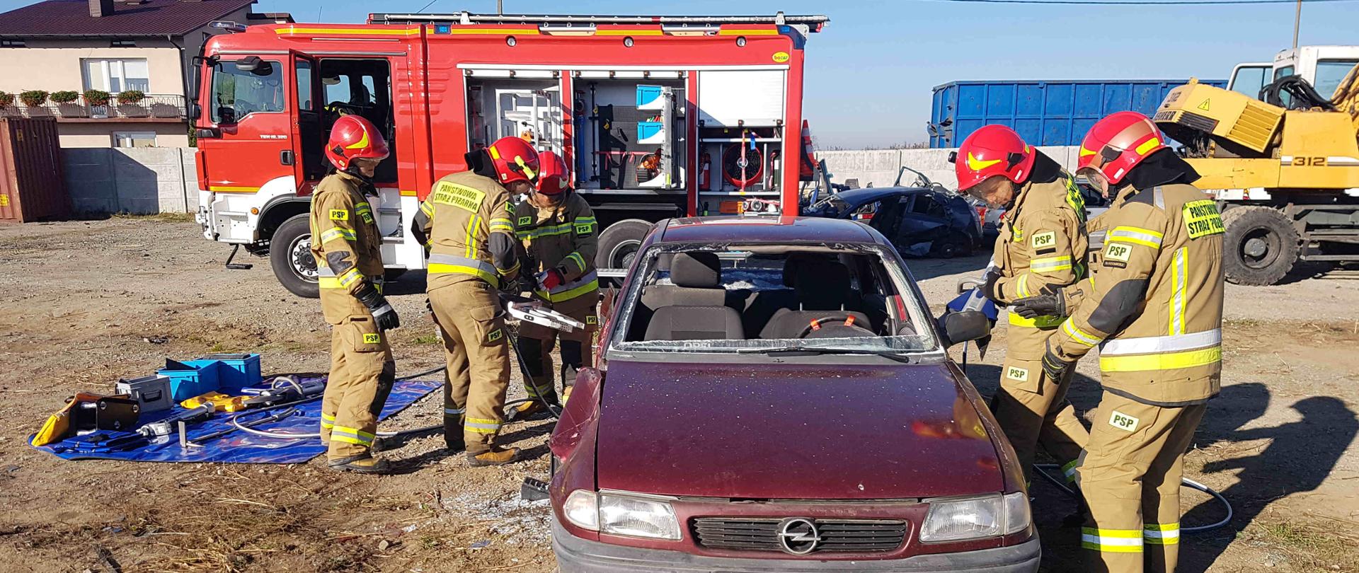 Zdjęcie zrobione w dzień na auto złomie. Na zdjęciu widać strażaków, którzy przy pomocy narzędzi hydraulicznych tną samochód osobowy. W oddali widać samochód strażacki. 