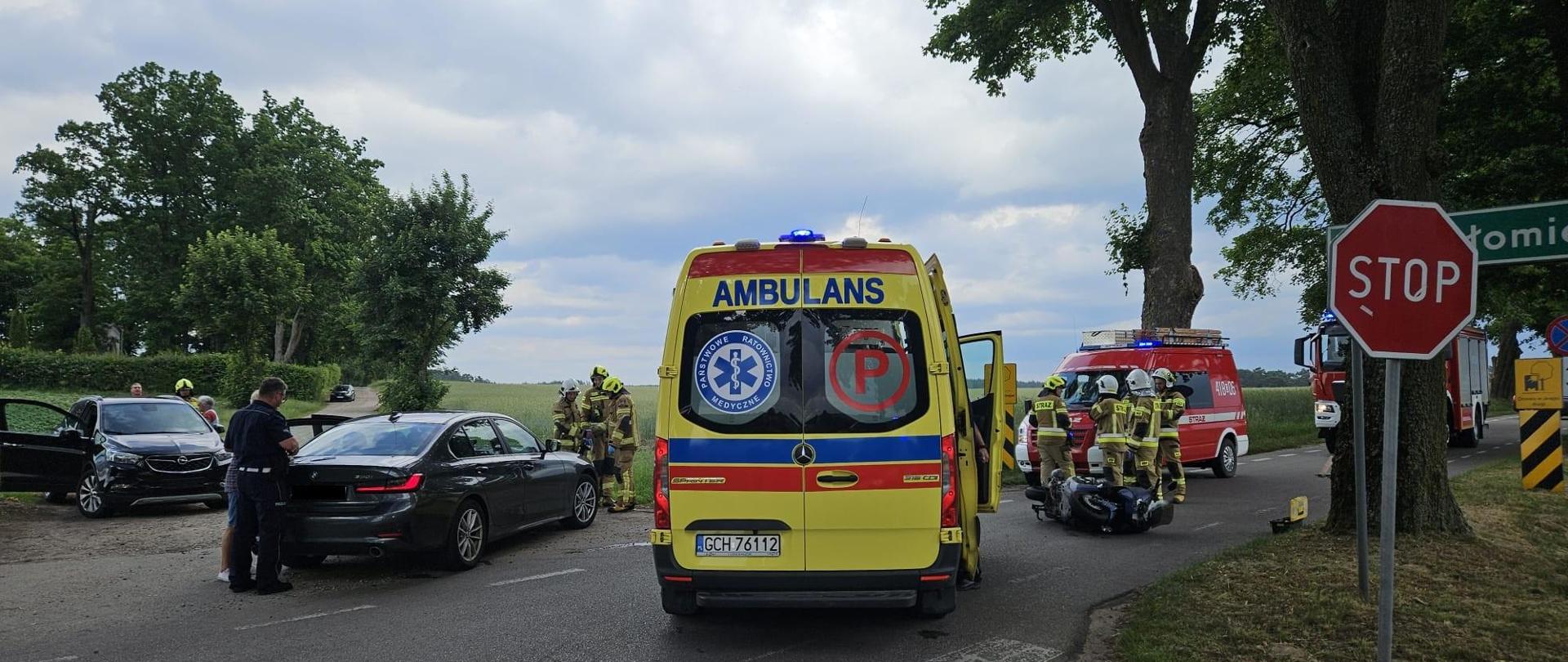 Pochmurny dzień, na pierwszym planie żółta karetka pogotowia ratunkowego z czerwonym napisem AMBULANS, karetka ma włączone niebieskie światła ostrzegawcze, stoi na środku drogi. Z prawej strony widać dwa czerwone samochody Straży Pożarnej na tle, których stoi czterech strażaków ubranych w piaskowe ubrania specjalne oraz białe i żółte hełmy, przed nimi leży motocykl. Z lewej strony na poboczu drogi stoją dwa czarne samochody, przy których stoją kilkuosobowe grupy ludzi. Pola wokół zdarzenia są intensywnie zielone.