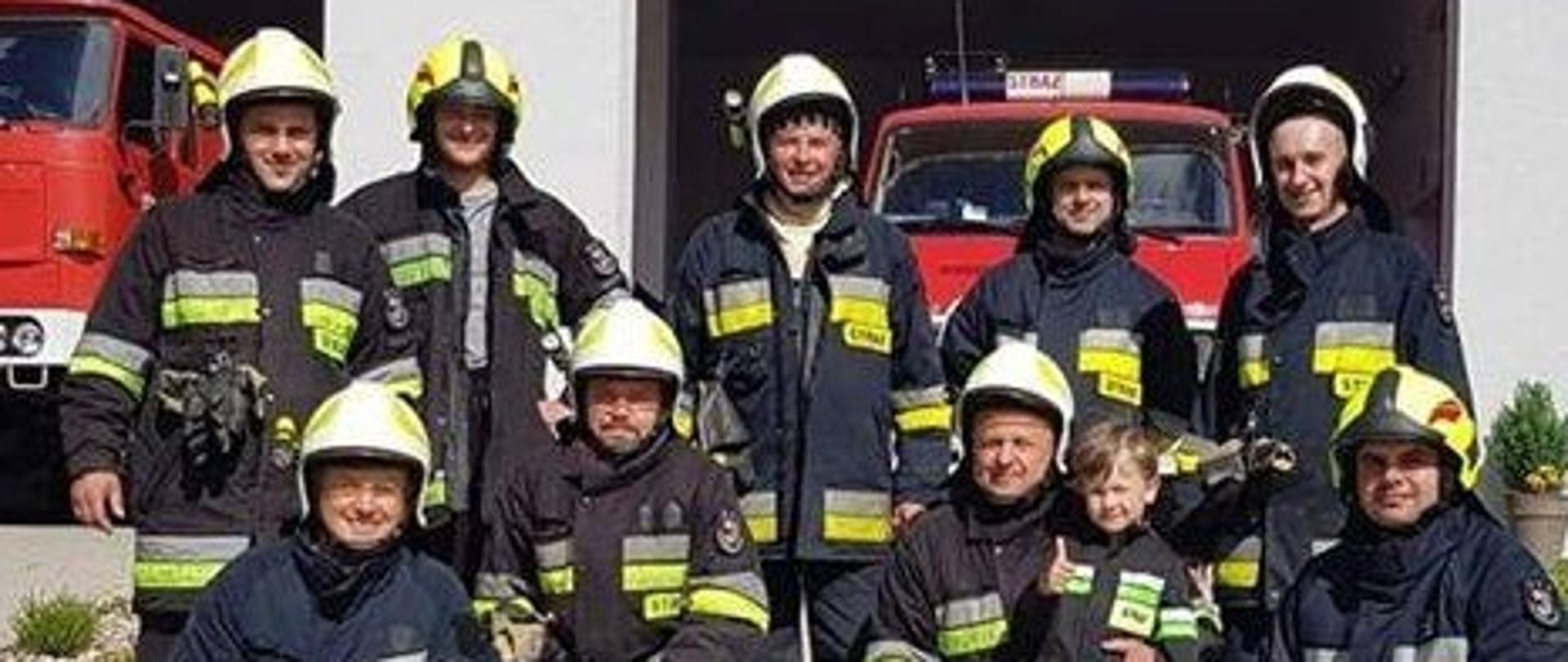 Zdjęcie przedstawia 9 ochotników ubranych w ubranie specjalne oraz białe hełmy. Jeden ze strażaków trzyma na kolanie chłopca ubranego w struj strażaka. W tle widać dwa boksy garażowe a w nich Dwa samochody strażackie.