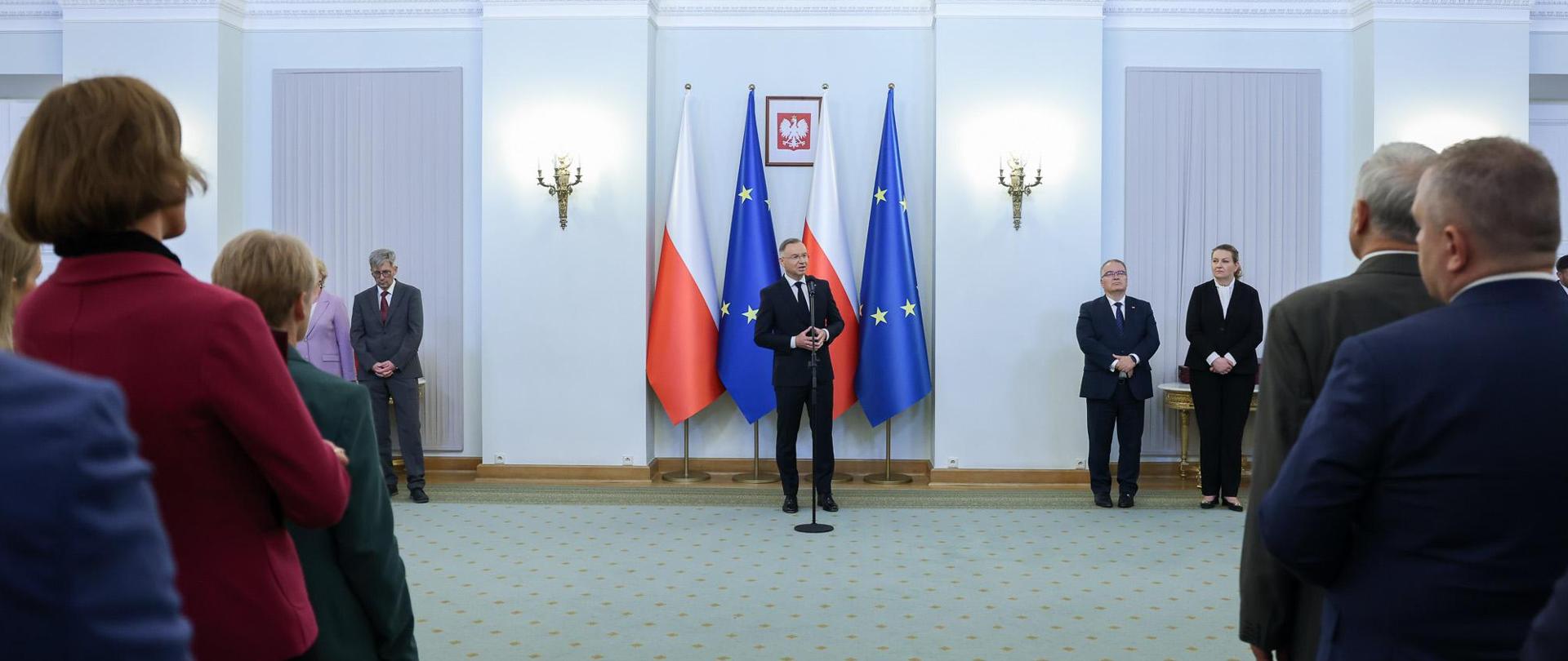 Widok z tyłu na dużą salę, w głębi prezydent Duda stoi i mówi do mikrofonu, za nim flagi Polski i UE, przed nim widać dużo ludzi.