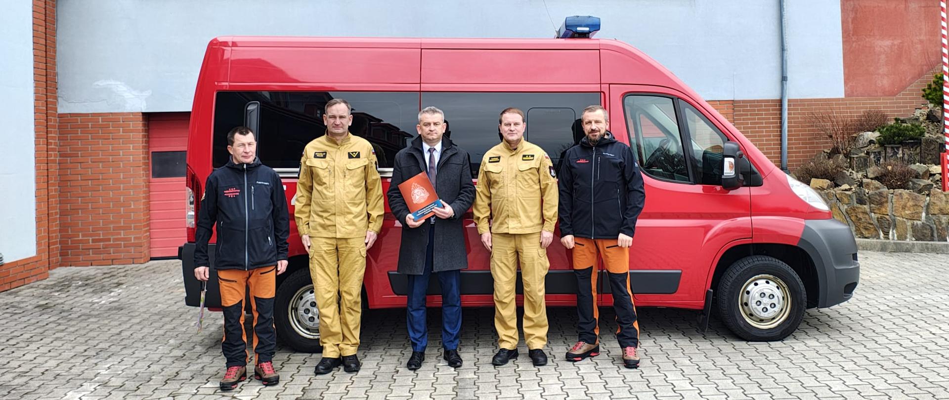 Zdjęcie grupowe wykonane na zewnątrz. W tle samochód oraz budynek. Na pierwszym planie 5 osób. 