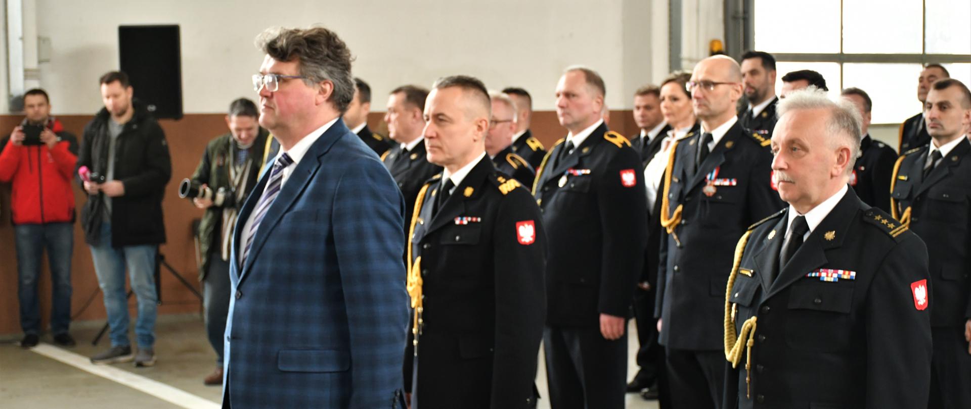 Na zdjęciu stoją na baczność strażacy w mundurach wyjściowych, z przodu przed nimi stoi Wiceminister Spraw Wewnętrznych i Administracji Pan Maciej Wąsik
