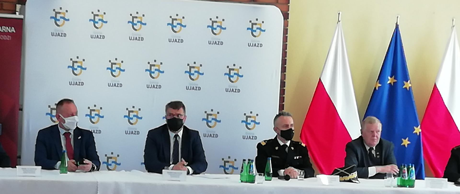 Czterech mężczyzn siedzi przy stole który przykryty jest białym obrusem. Jeden to strażak. Za siedzącymi w centralnej części znajduje się baner z logo Gminy Ujazd a po prawej stronie trzy flagi. Dwie skrajne to flagi biało-czerwone natomiast środkowa to flaga Unii Europejskiej. 