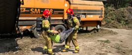 Dwóch strażaków ubranych w sprzęt ochrony dróg oddechowych ewakuuje pracownika zakładu. 