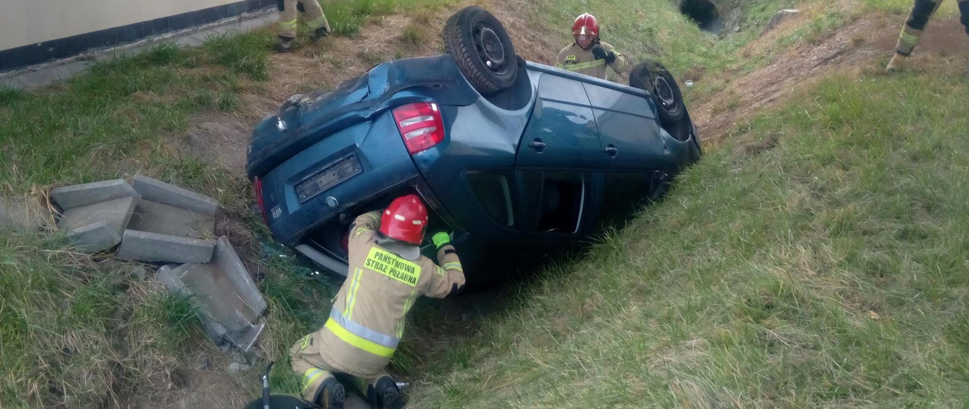 Zdjęcie przedstawia samochód osobowy leżący w pozycji na dachu w przydrożnym rowie i pracujących przy nim ratowników.