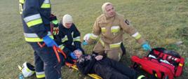 Teren stadionu w Rawiczu. Na trawie , na noszach typu deska lezy "poszkodowana" osoba, z zakrwawiona twarzą. Trzech strażaków, uczestników warsztatów udziela jej kwalifikowanej pierwszej pomocy. Występują w ubraniach specjalnych. Obok znajduje się torba ze sprzętem medycznym.