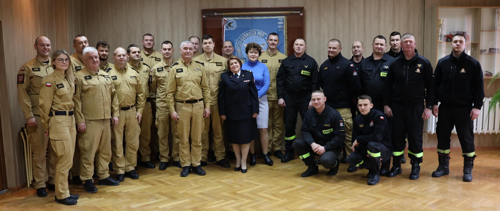 Przejście na zaopatrzenie emerytalne bryg. Podporska Hanna - strażacy stoją w grupie do zdjęcia pamiątkowego w świetlicy, w środku emerytka oraz Komendant, w tle sztandar KM PSP Ostrołęka.