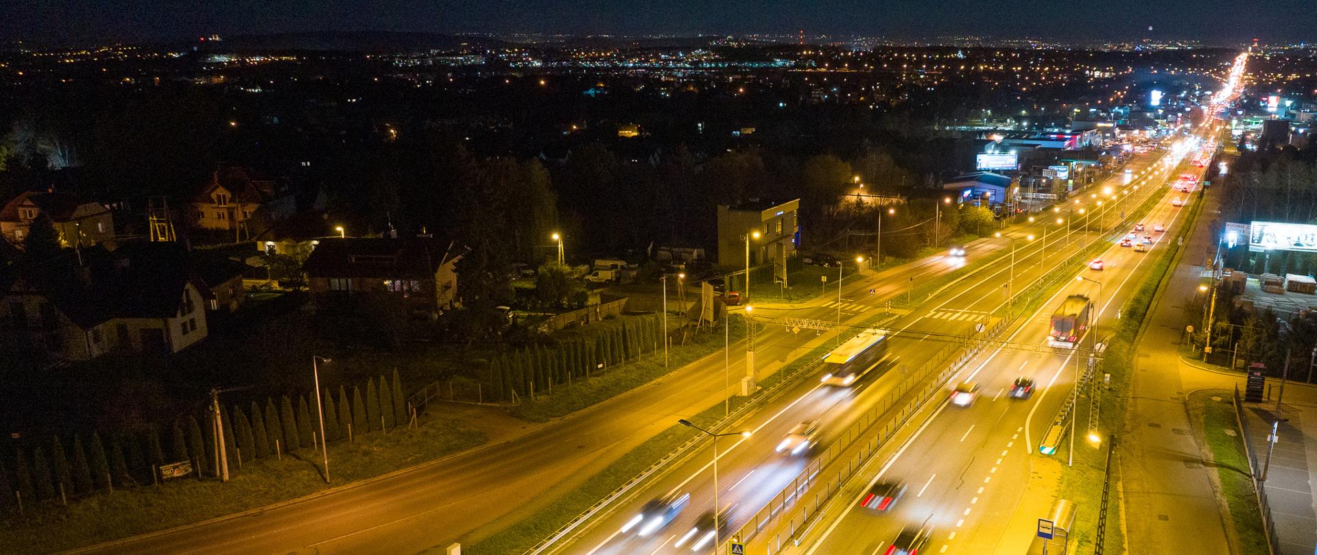Zdjęcie przedstawia drogę krajową nocą. Droga ma po dwa pasy ruchu w obie strony. Jest dobrze oświetlona. Poruszają się po niej różnego rodzaju pojazdy - samochody osobowe, autobusy. W tle widoczna nocna panorama miasta z licznymi światłami.