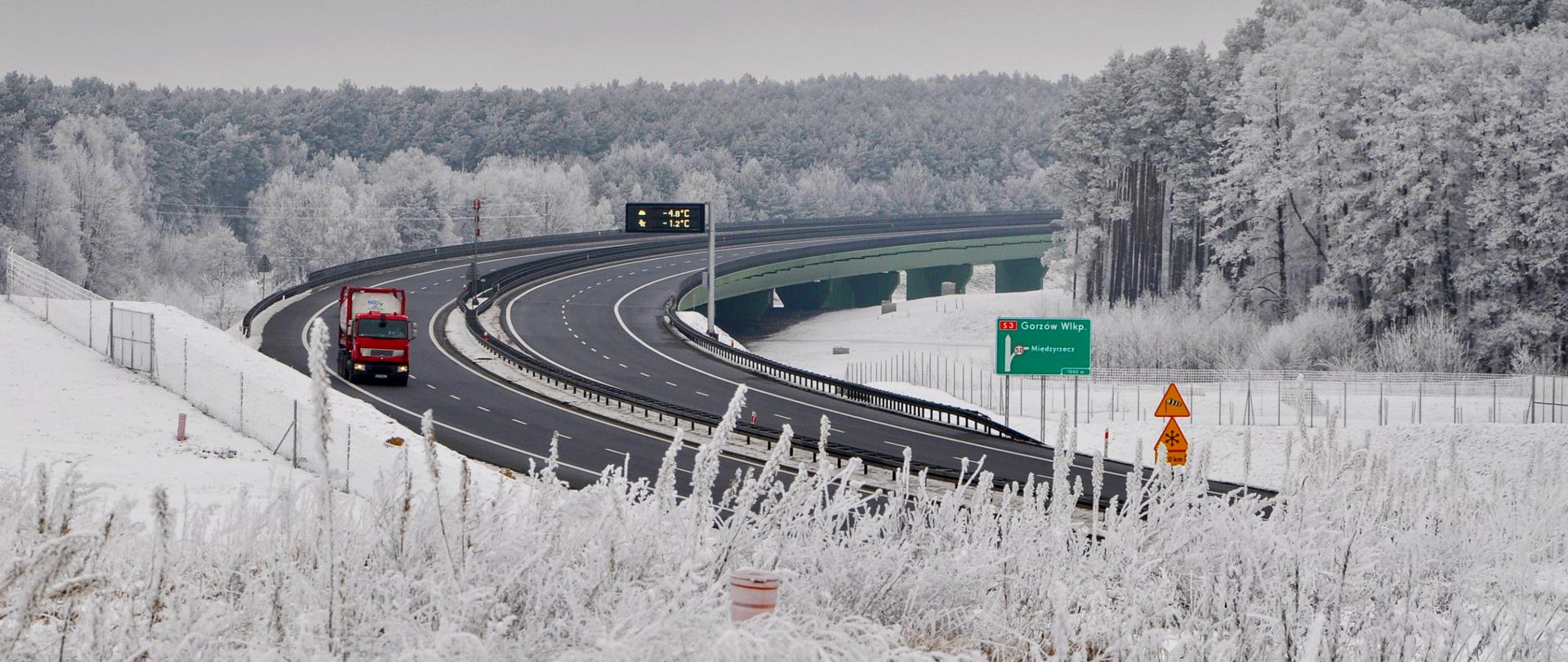 Zdjęcie przedstawia drogę ekspresową S3 w zimowej scenerii. Droga jest czarna i odśnieżona. Teren przy drodze jest pokryty śniegiem, a otaczające ją drzewa są oszronione. Na drodze widoczna jest czerwona ciężarówka, a w tle znajduje się most. Po prawej stronie drogi znajduje się zielony znak drogowy wskazujący kierunki do Gorzowa Wielkopolskiego i Międzyrzecza. Na tablicy informacyjnej nad drogą widnieje temperatura: -4.8°C oraz -1.2°C.