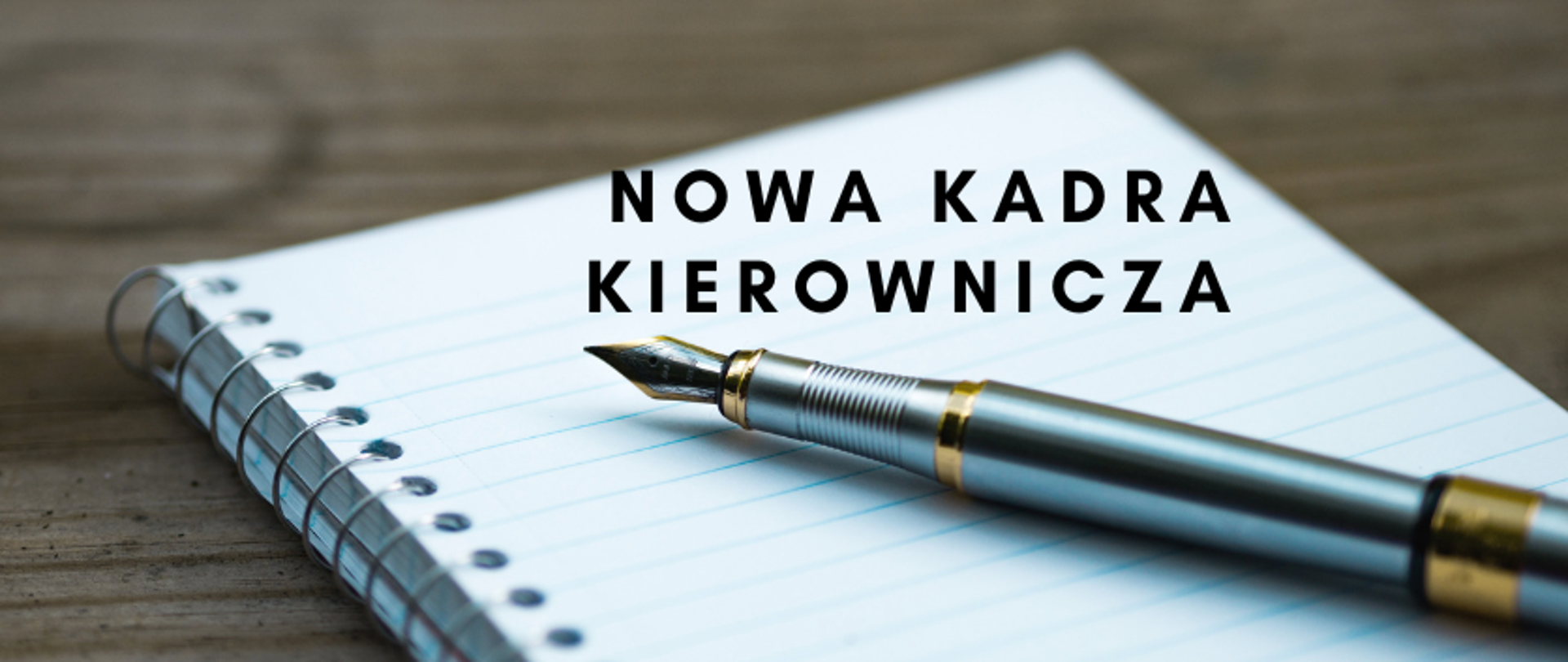 grafika: na drewnianym blacie leży rozłożony kołonotes, a na nim jest położone pióro do pisania. Napis: nowa kadra kierownicza