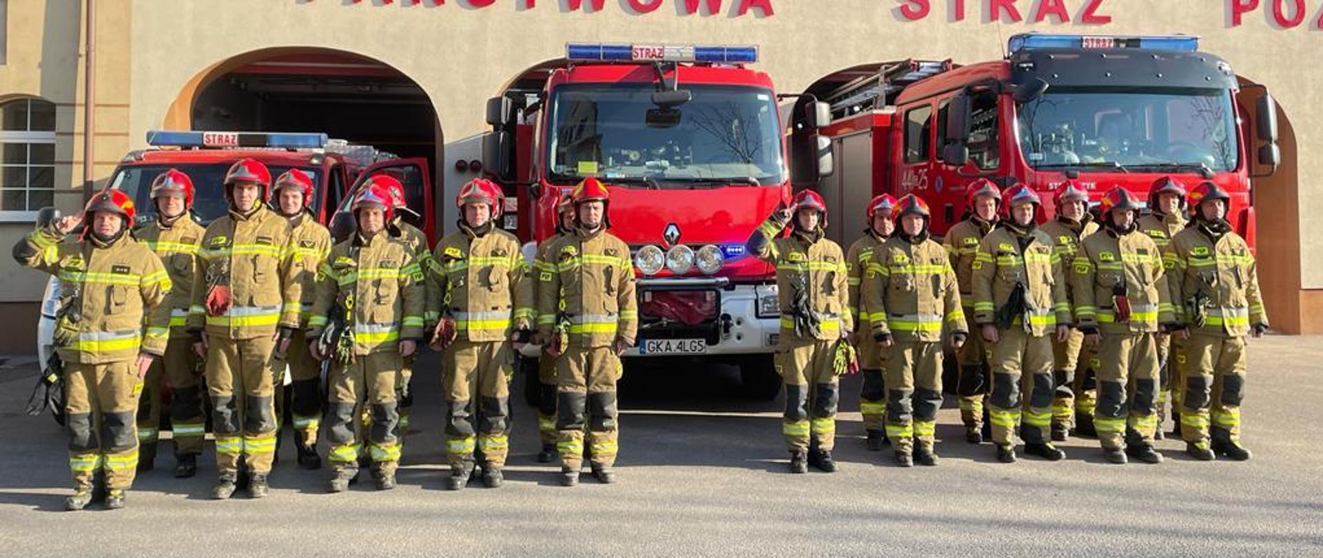 Strażacy ustawieni w dwuszeregu na tle samochodów pożarniczych ustawionych przed budynkiem komendy.