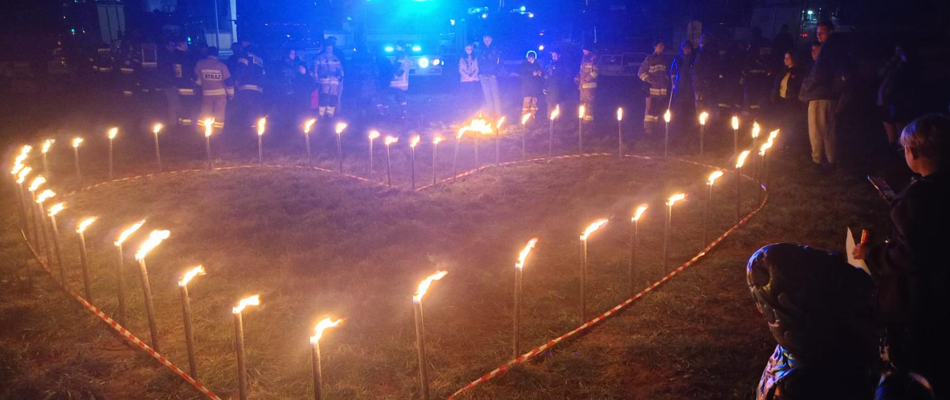 Na zdjęciu jest wieczór, na trawniku serce z zapalonych pochodni wokół niego ustawieni ludzie.