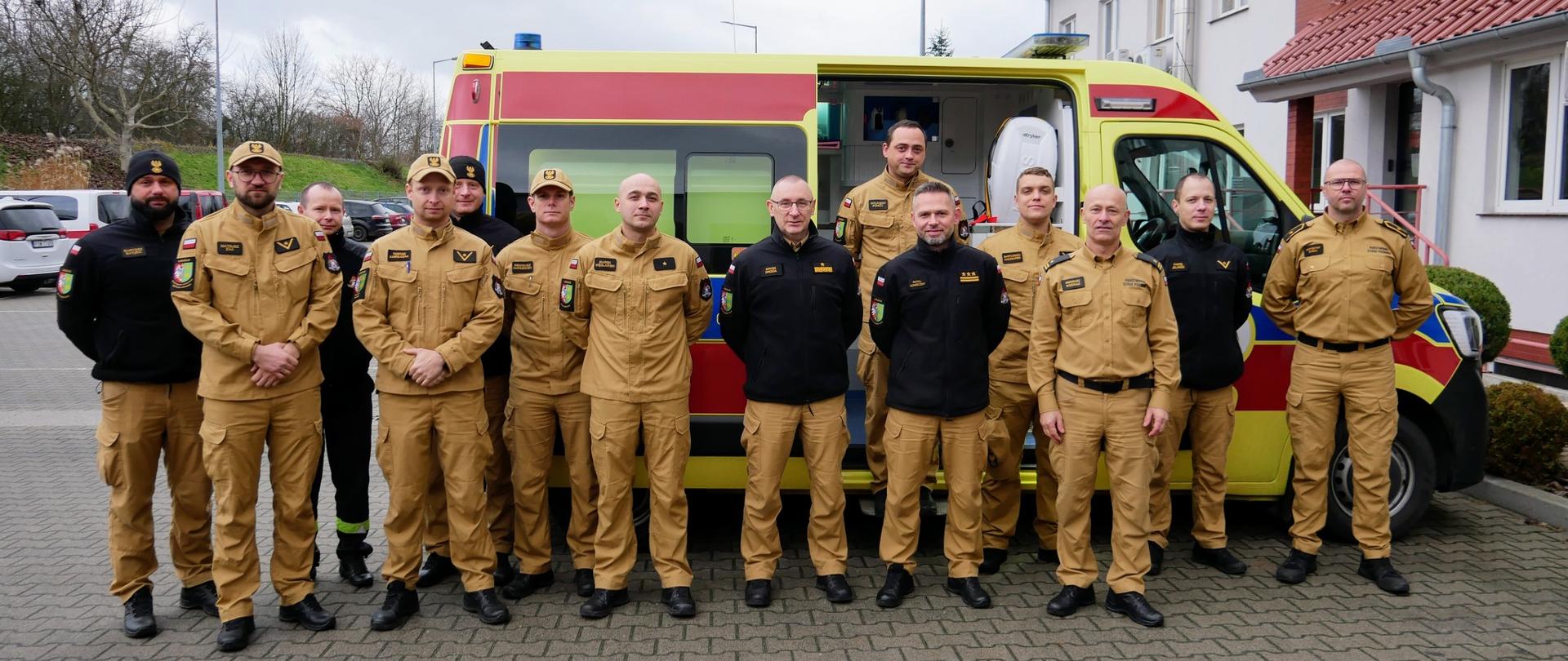 Zdjęcie przedstawia grupę ratowników PSP w musztardowych mundurach, stojących przed kolorowym ambulansem na placu przed budynkiem. Wszyscy członkowie zespołu patrzą w stronę obiektywu, mają naszywki i odznaczenia na mundurach, a w tle widać budynek z czerwonym dachem.