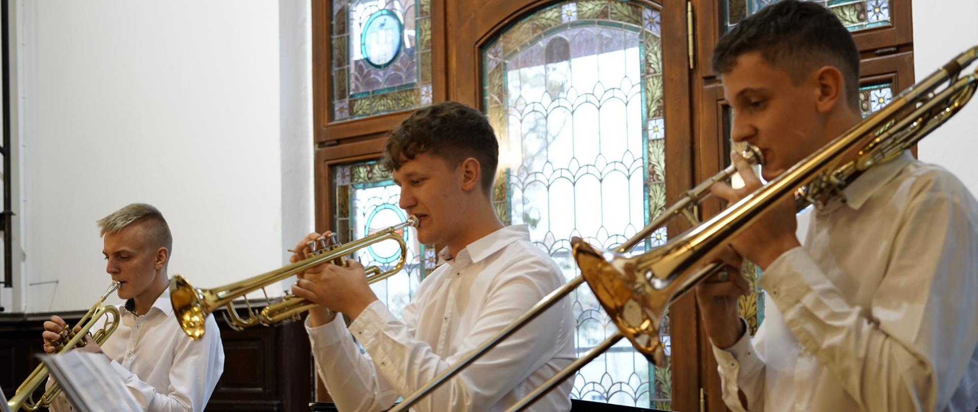 Koncert muzyczny sekcji dętej. Trzech chłopaków wieku nastoletnim gra kolejno od prawej: na puzonie, na trąbce i na trąbce. Patrzą na nuty ułożone na pulpicie. Są ubrani w białe koszule. Za nimi, w tle zdjęcia znajduje się ogromne okno pełne witraży.