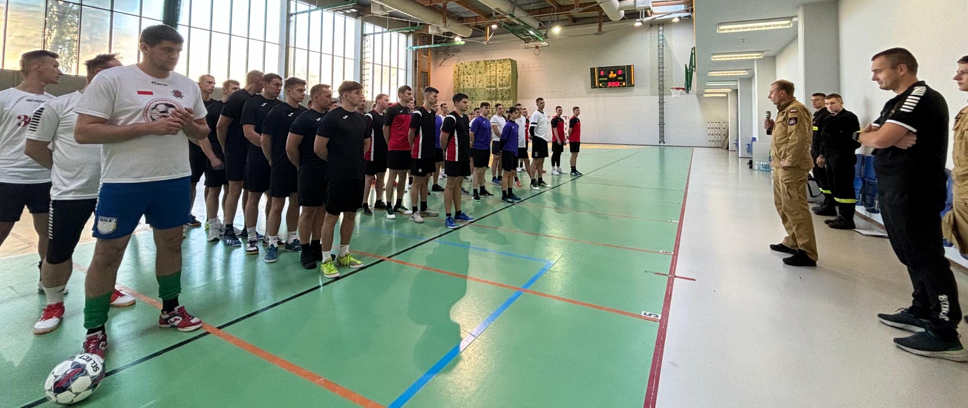 Hala sportowa SA PSP w Krakowie. Rozpoczęcie szkolnej ligi futsal. Po prawej stronie ustawienie w rzędach (gęsiego) zawodnicy poszczególnych drużyn, po prawej otwierający wydarzenie z-ca komendanta SA PSP w Krakowie mł.bryg. Dariusz Ruchała kadeci oraz organizatorzy. Mężczyźni w strażackich mundurach i strojach sportowych.