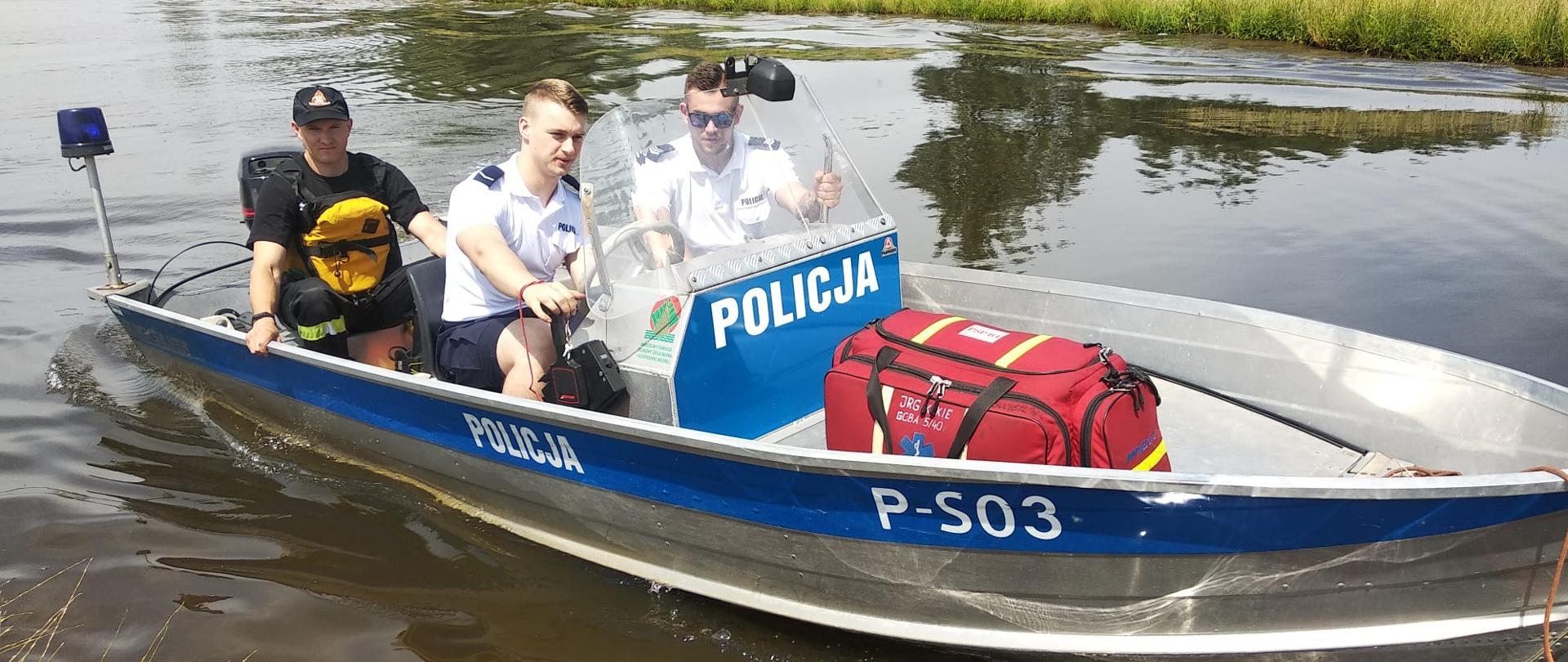 Policyjna łódka na zalewie "Antoniów" blisko nabrzeża, na pokładzie dwóch policjantów i strażak ze sprzętem do ratownictwa wodnego