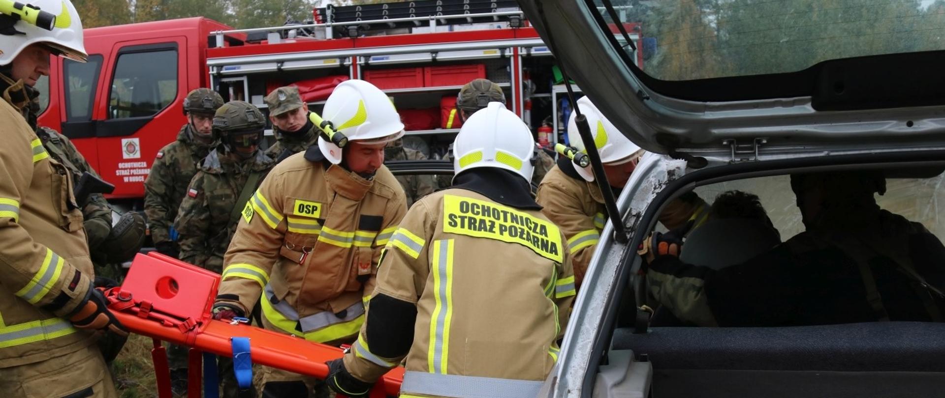 Zdjęcie przedstawia czterech strażaków ochotników podczas ewakuacji poszkodowanego z samochodu przy użyciu deski ortopedycznej. W tle żołnierze WOT i samochód ratowniczo-gaśniczy straży pożarnej 