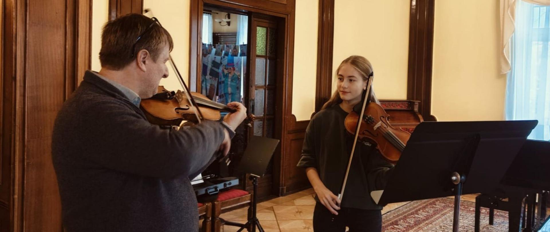 Zdjęcie kolorowe. Jasnowłosa uczennica ze skrzypcami obserwuje Pana grającego na skrzypcach.