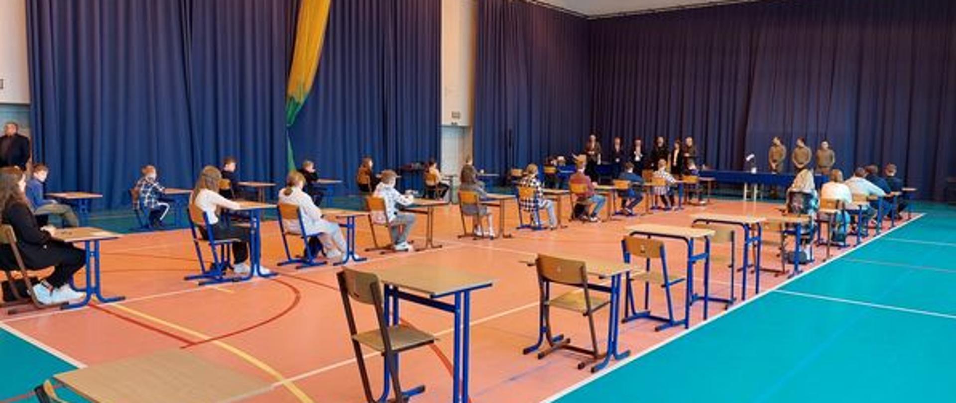 Uczniowie siedzą przy pojedynczych ławkach ustawionych w trzech rzędach na sali gimnastycznej. Przed nimi stoją organizatorzy i komisja. 