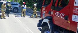 Na zdjęciu widać pojazd ratownniczy i strażaków podczas akcji, na drodze samochód po wypadku blokuje dwa pasy ruchu w oddali funkcjonariusze Policji.