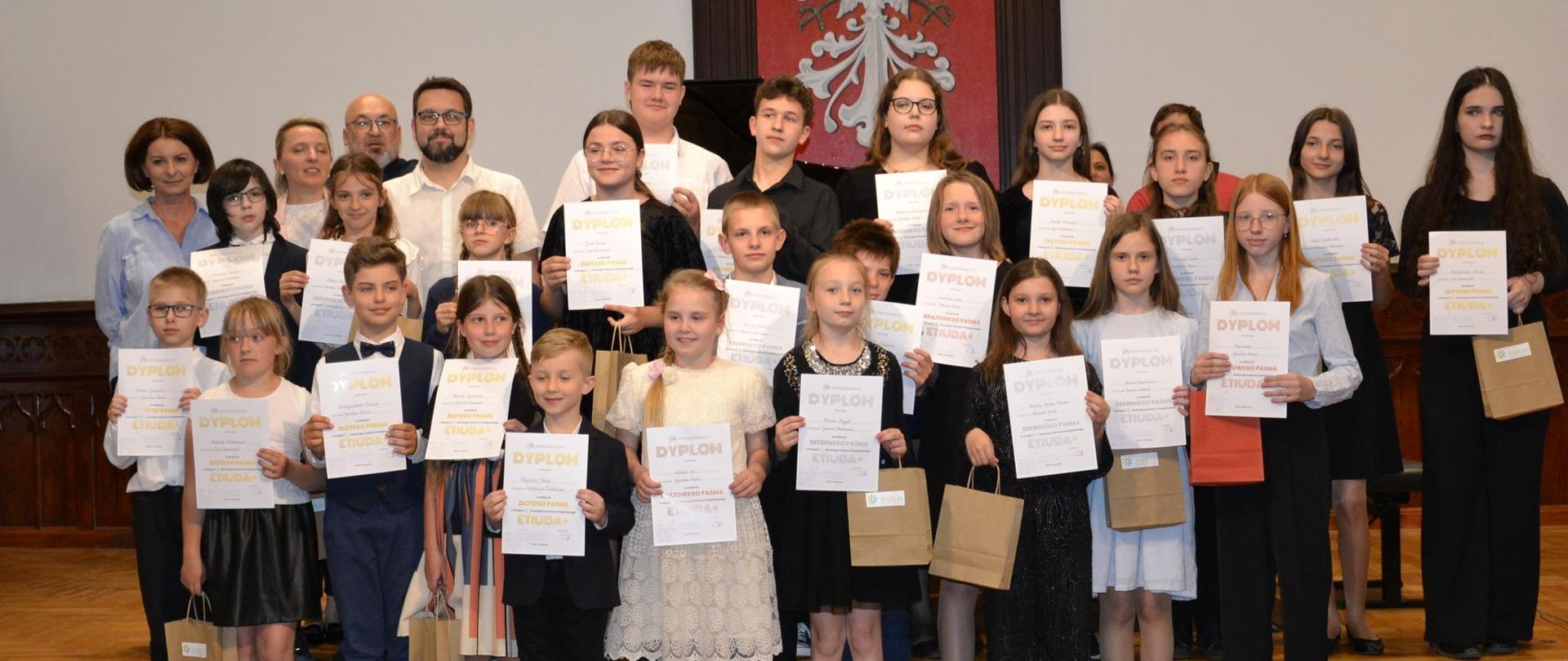 uczestnicy szkolnego konkursu fortepianowego Etiuda+ trzymający dyplomy i nagrody wraz z nauczycielami na scenie w Sali Królewskiej PSM Mielec