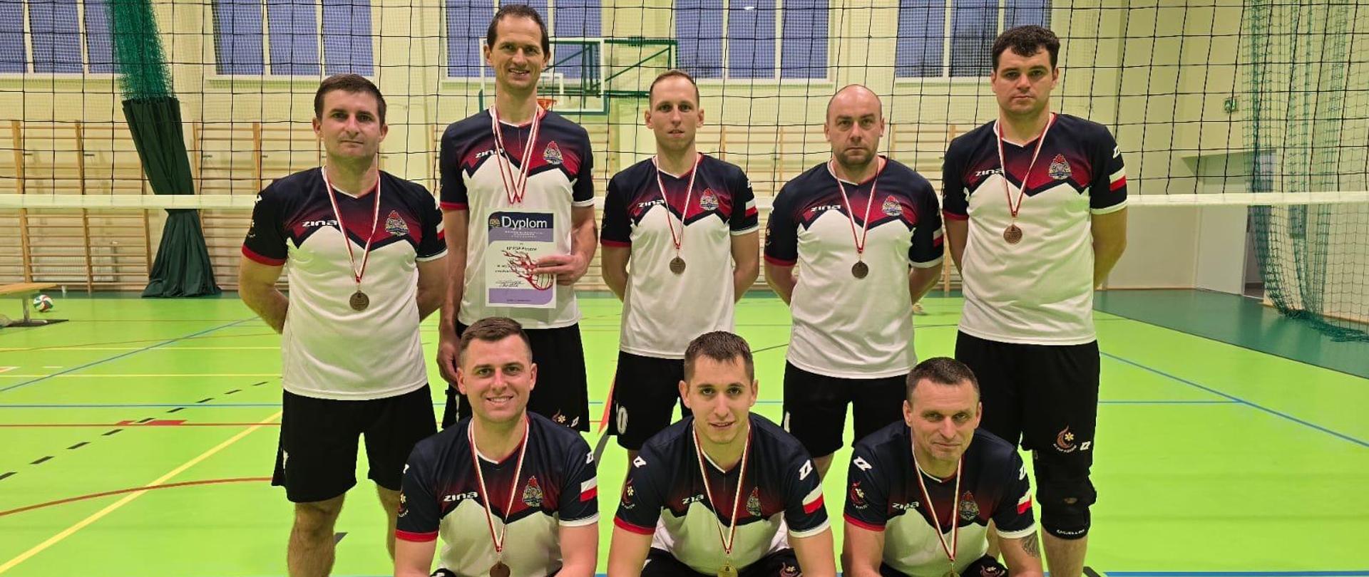 Zdjęcie grupowe zawodników, na sali sportowej ośmiu mężczyzn w strojach sportowych ustawionych w dwuszeregu, pierwszy szereg klęczy, przed nimi widoczny jest puchar