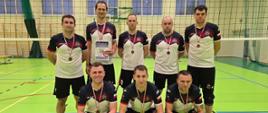 Zdjęcie grupowe zawodników, na sali sportowej ośmiu mężczyzn w strojach sportowych ustawionych w dwuszeregu, pierwszy szereg klęczy, przed nimi widoczny jest puchar