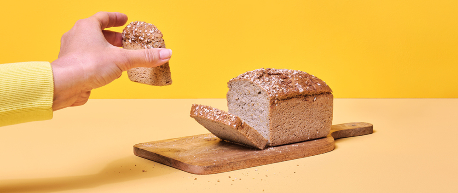 Na zdjęciu znajduje się bochenek chleba na desce do krojenia. Po lewej stronie widoczna jest dłoń, w której znajduje się kromka chleba. Tło jest żółte.