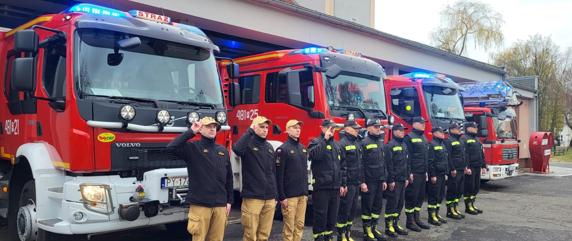 Strażacy oddali hołd tragicznie zmarłemu policjantowi. Na zdjęciu widać strażaków przed budynkiem Komendy, samochody strażackie wyjechały z garażu, mają włączone sygnały świetlne i dźwiękowe, widać maszt z flagą Polski i budynek Komendy Powiatowej PSP w Kościanie.