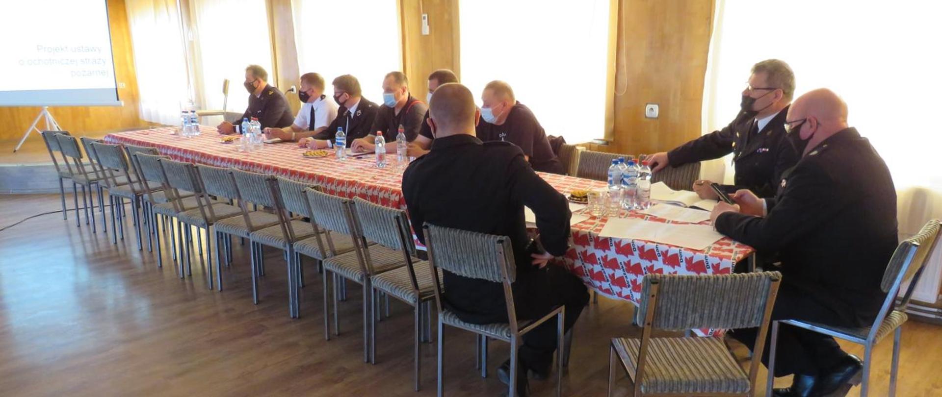 strażacy OSP i PSP w mundurach wyjściowych i maseczkach, siedzą za stołem i słuchają 