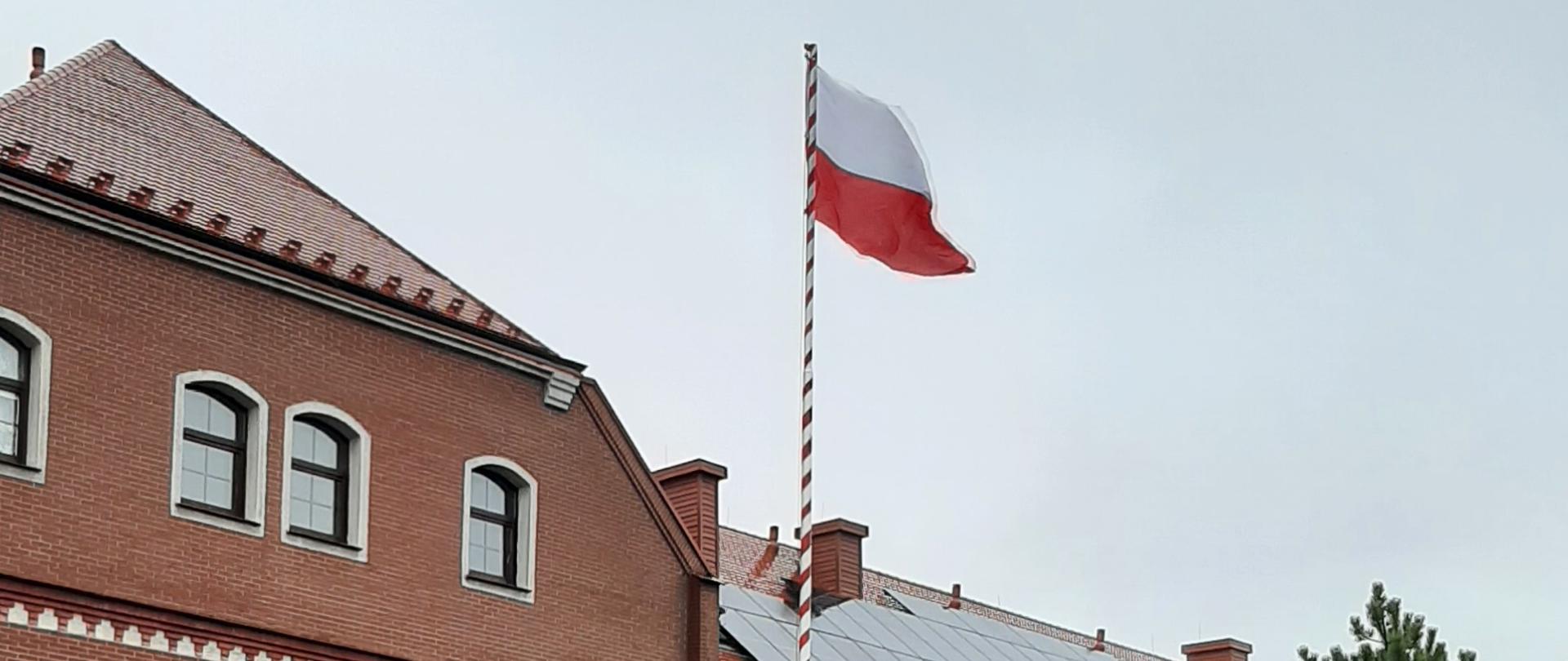 Zdjęcie przedstawia biało czerwoną flagę na maszcie. W głębi zdjęcia widać budynek KP PSP Prudnik.