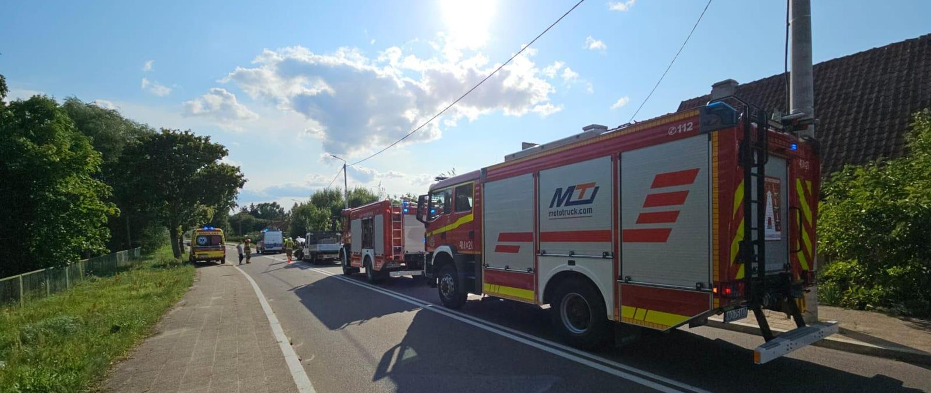 Na drodze na prawym pasie stoją dwa samochody strażackie. Dalej w oddali samochód dostawczy. Na lewym pasie karetka pogotowia. Słonecznie, niebieskie niebo.