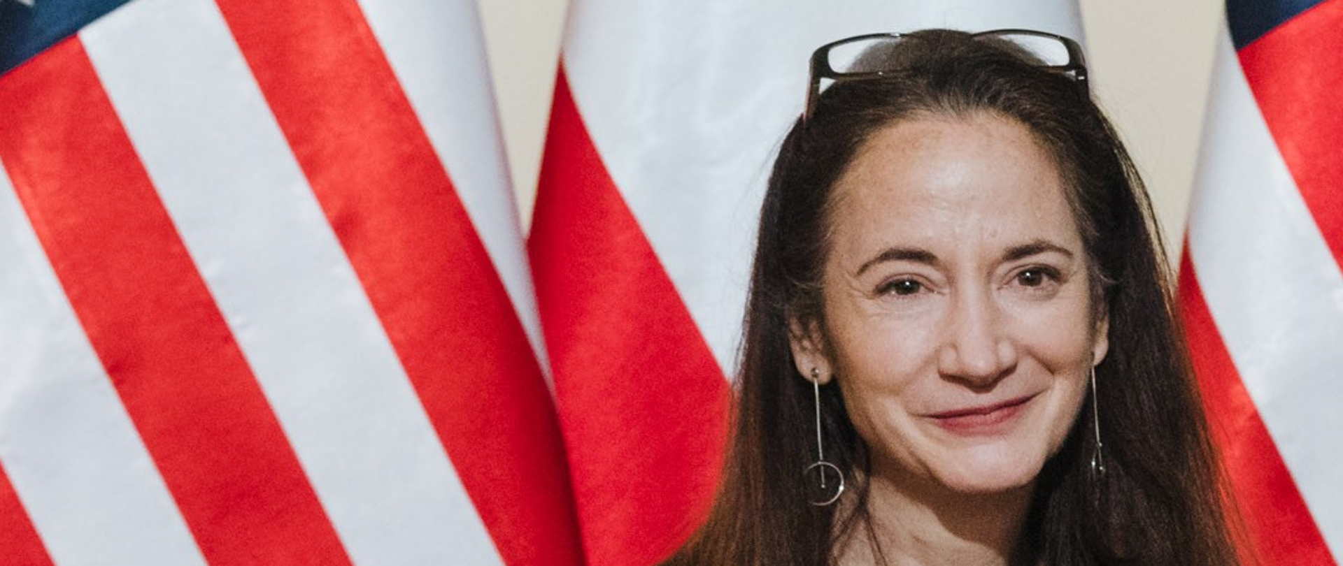 Portret uśmiechniętej kobiety z długimi rozpuszczonymi ciemnymi włosami na tle amerykańskiej i polskiej flagi